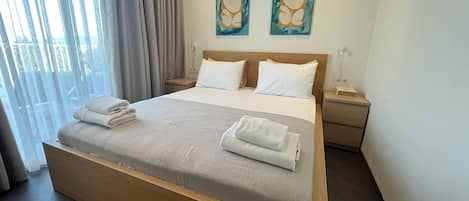 A neatly made bed with white linens and towels, a wooden headboard, two wall-mounted paintings, a bedside table with a lamp, and a view through a window with curtains.