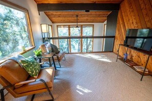 Una sala de estar acogedora con un sofá, sillas, una mesa de centro y un televisor sobre un mueble de madera. Amplios ventanales ofrecen vistas al exterior.