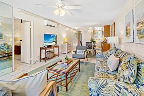 A living room with a sofa, coffee table, and a television. There is a dining area with a table and chairs. The room has a ceiling fan and a window.