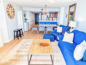 Ein Wohnzimmer mit blauem Sofa, Couchtisch aus Holz und Essbereich mit weißen Stühlen.