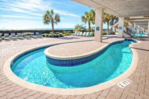 A swimming pool with a tiled edge, lounge chairs, palm trees, and a view of the ocean.