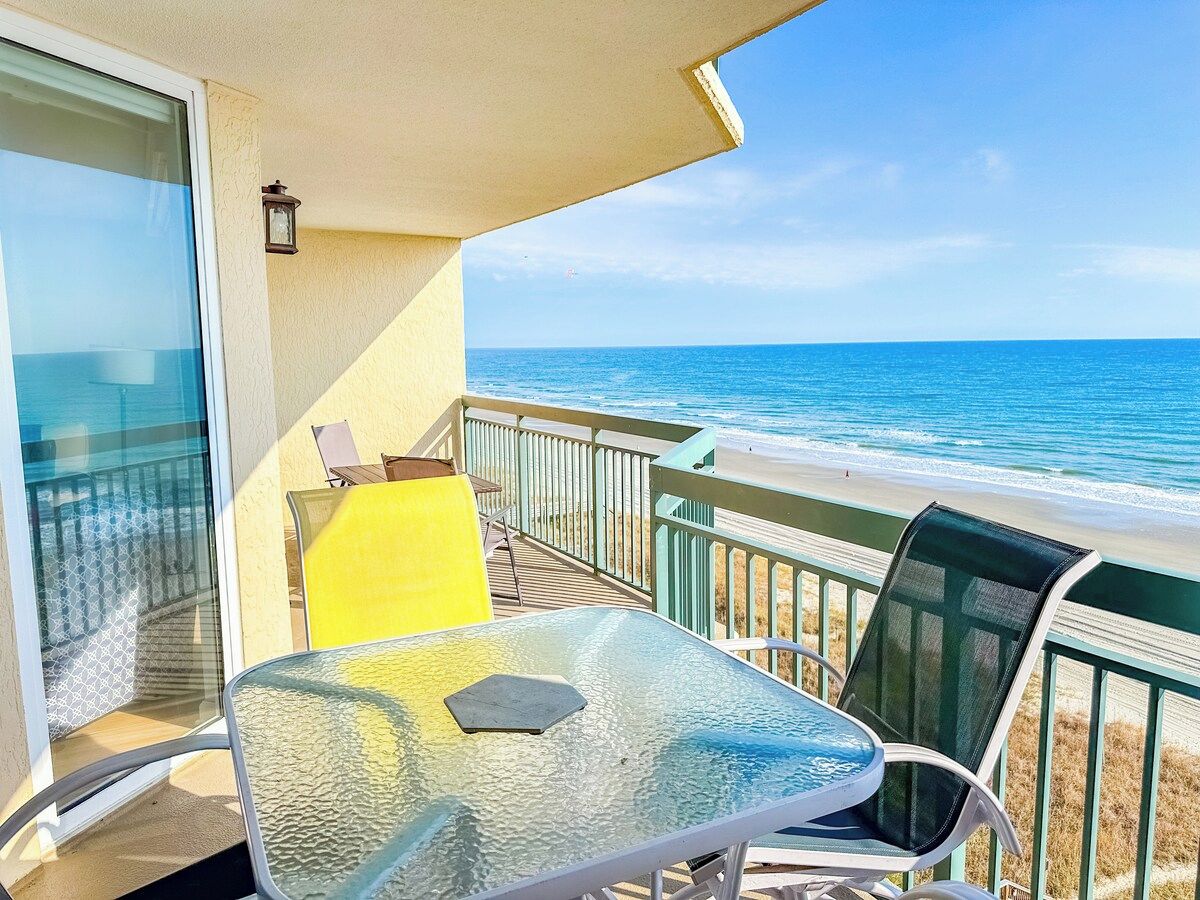 A balcony with a glass table and chairs, overlooking a beach with the ocean in the background.