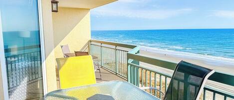 A balcony with a glass table and chairs, overlooking a beach with the ocean in the background.