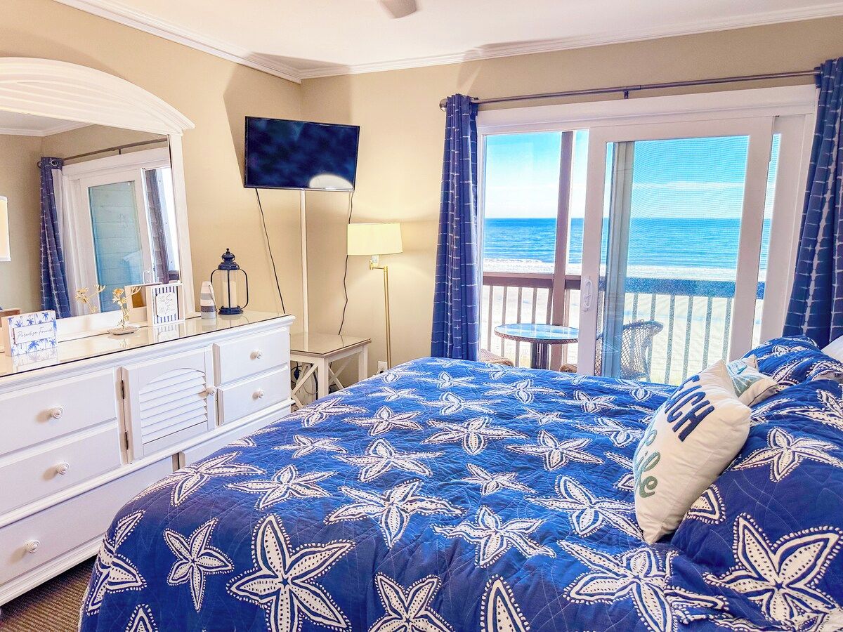 A bedroom with a large bed, a dresser, a television, and a view of the ocean through a balcony.