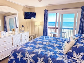 A bedroom with a large bed, a dresser, a television, and a view of the ocean through a balcony.