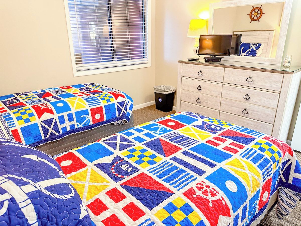 Une chambre avec deux lits, une commode, un téléviseur et une fenêtre avec des stores.