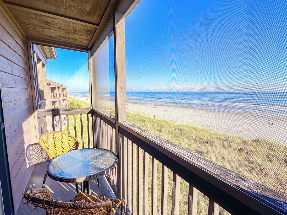 A balcony with a view of the beach, a table, and a chair.