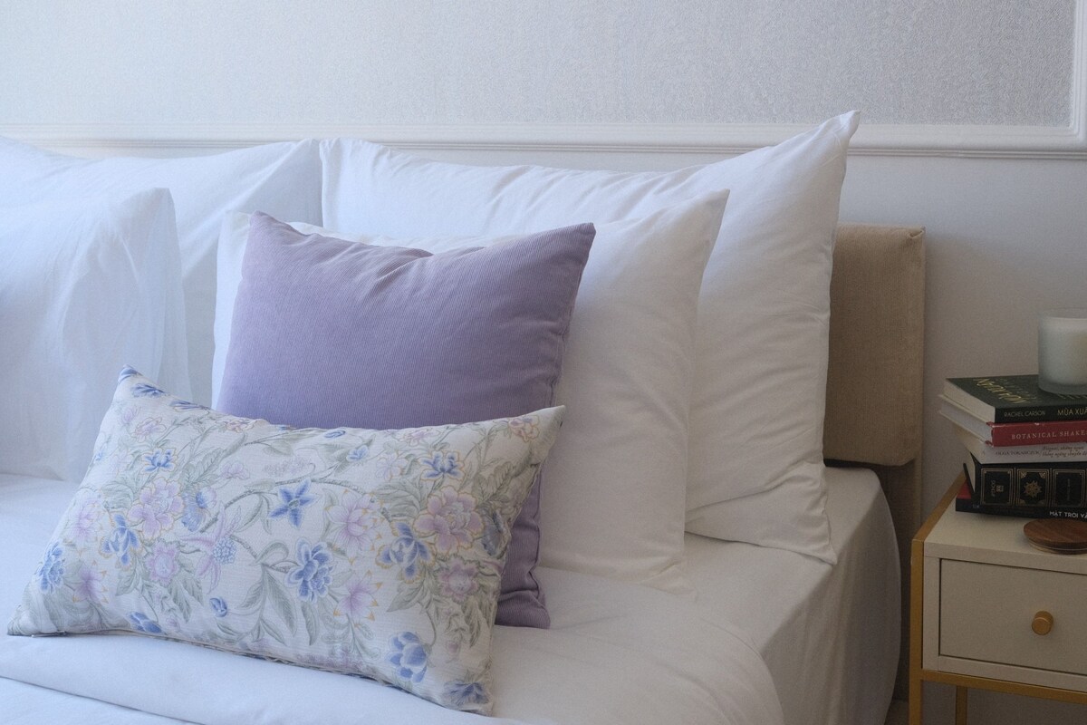 Una cama bien hecha con almohadas blancas y moradas, una almohada estampada con motivos florales y una mesita de noche con libros y una vela.