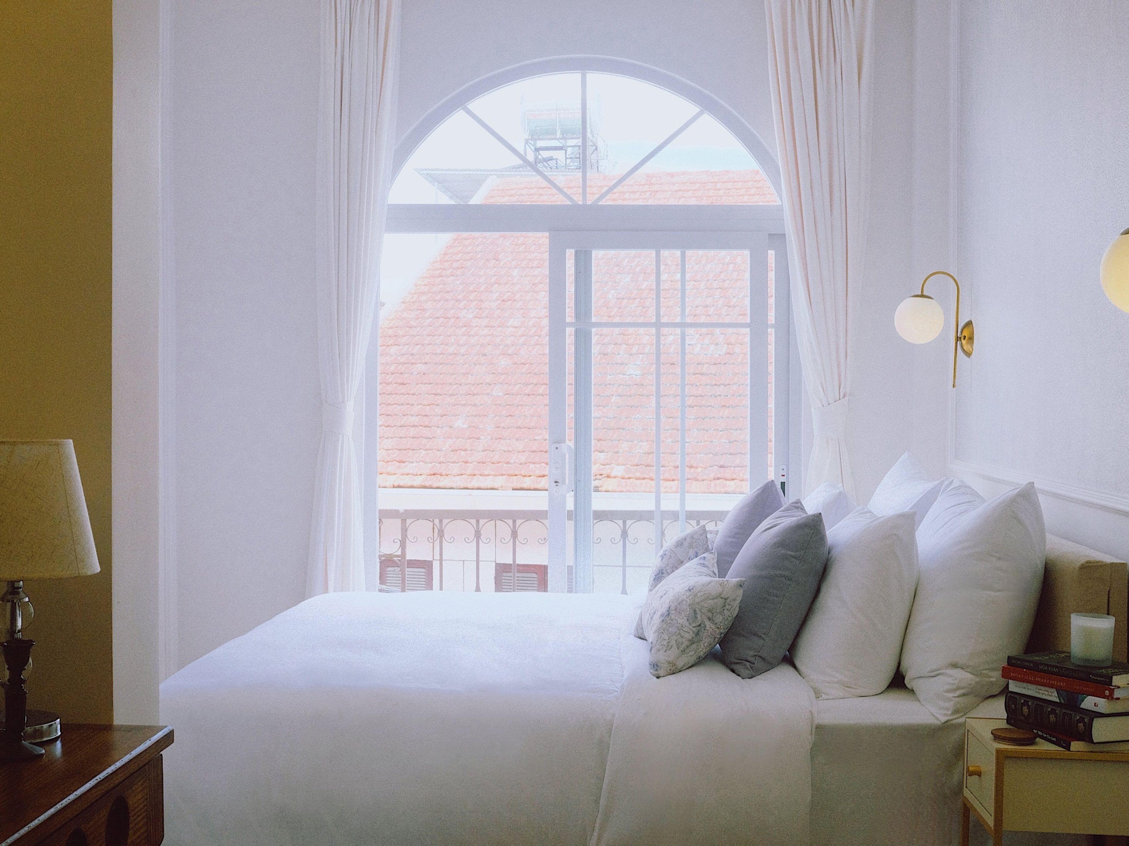 A bedroom with a large arched window, a bed with pillows, a bedside table with a lamp, and a wall-mounted light fixture.
