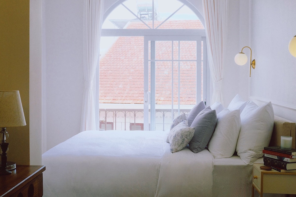 A bedroom with a large arched window, a bed with pillows, a bedside table with a lamp, and a wall-mounted light fixture.