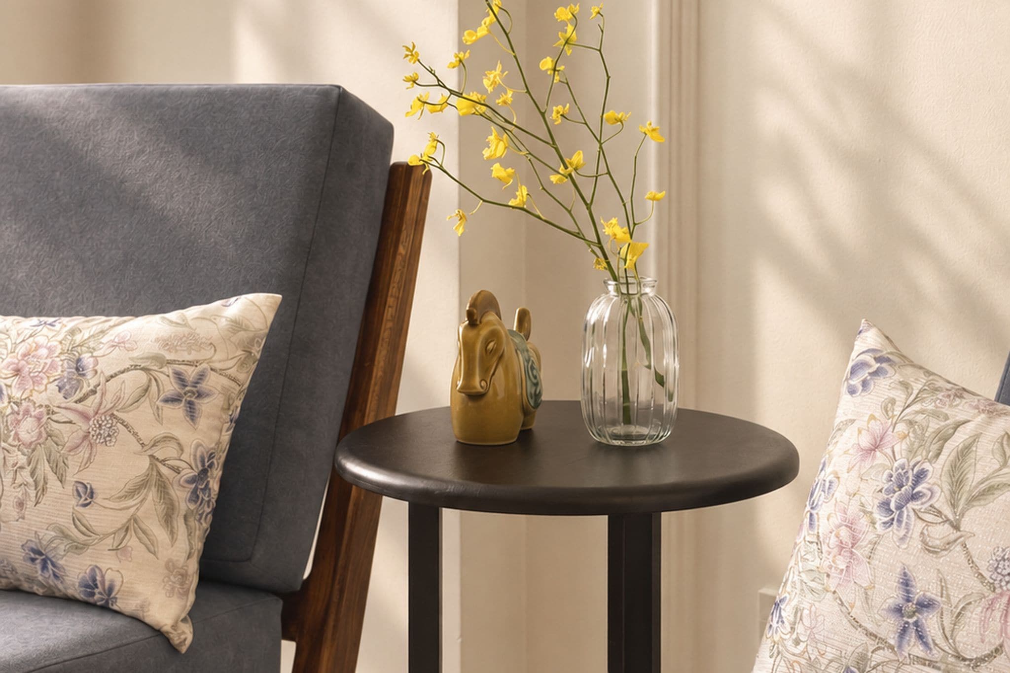 A room with a grey sofa, a small round table with a vase of yellow flowers, and a decorative horse figurine.