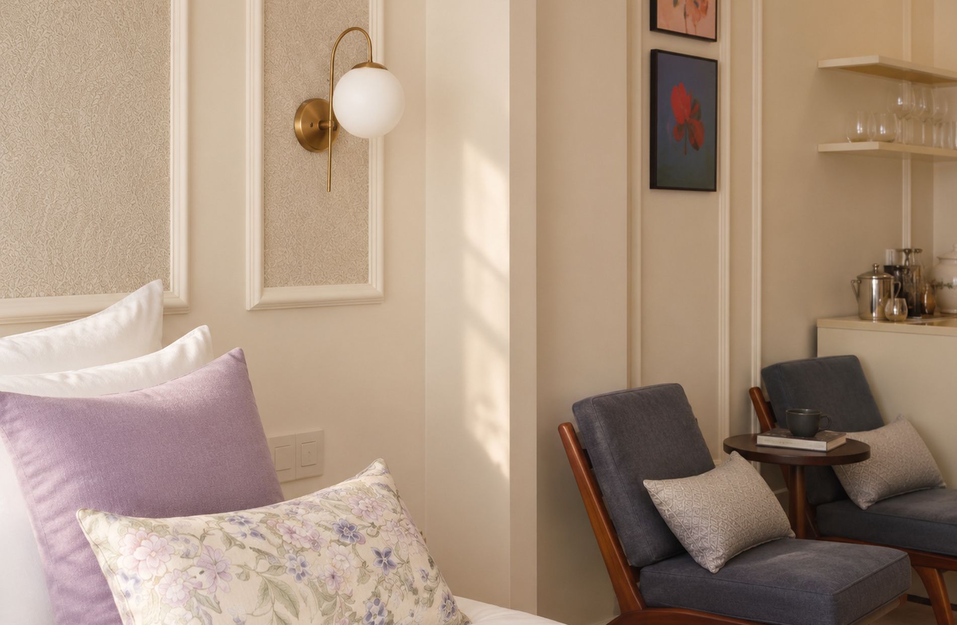 A room with a bed, two chairs, a small table, a wall-mounted light fixture, and a shelf with glassware.