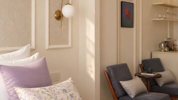 A room with a bed, two chairs, a small table, a wall-mounted light fixture, and a shelf with glassware.