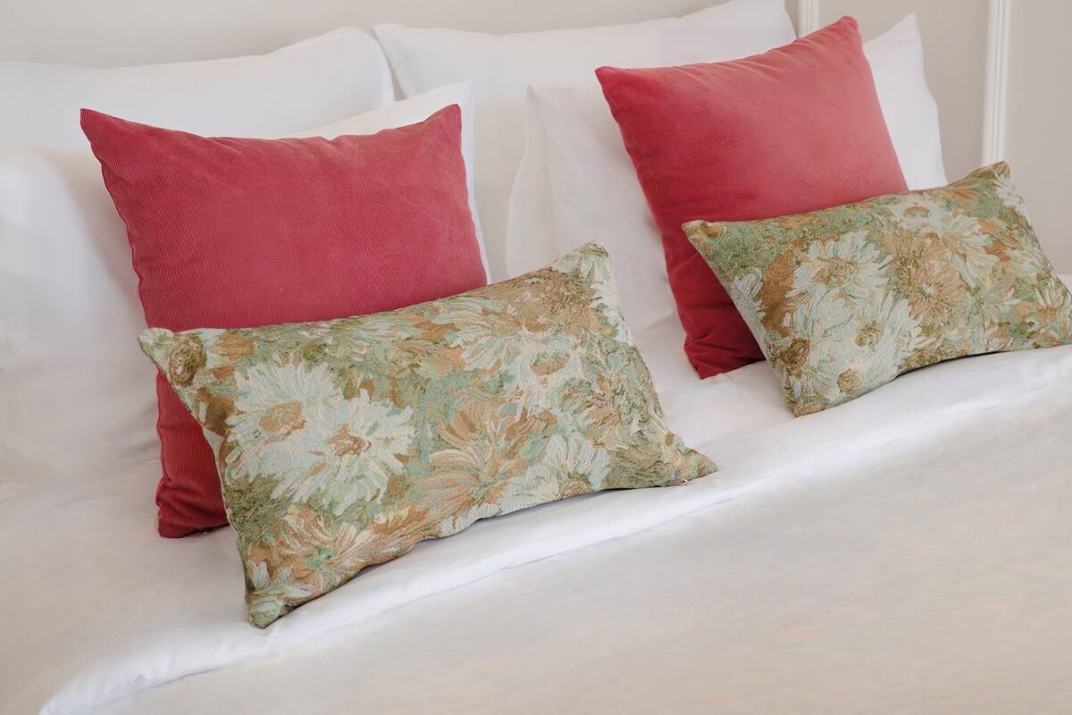 Una cama con ropa blanca, dos almohadas rojas y dos almohadas con estampado floral.