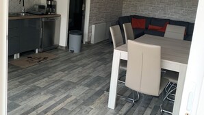 A modern kitchen with grey cabinets, a built-in dishwasher, and a coffee maker. There is a dining area with a white table and chairs, and a grey and red sofa. The floor is wooden with a herringbone pattern. - Appartement Proche Jonzac (Meux)