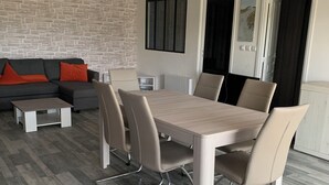 A modern dining area with a wooden table and chairs, a stone wall, and a sofa with orange cushions. - Appartement Proche Jonzac (Meux)