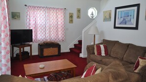 A living room with a television, a sofa, a coffee table, a staircase, and artwork on the walls. - Finisterre – Your Topsail Island Escape (North Topsail Beach)