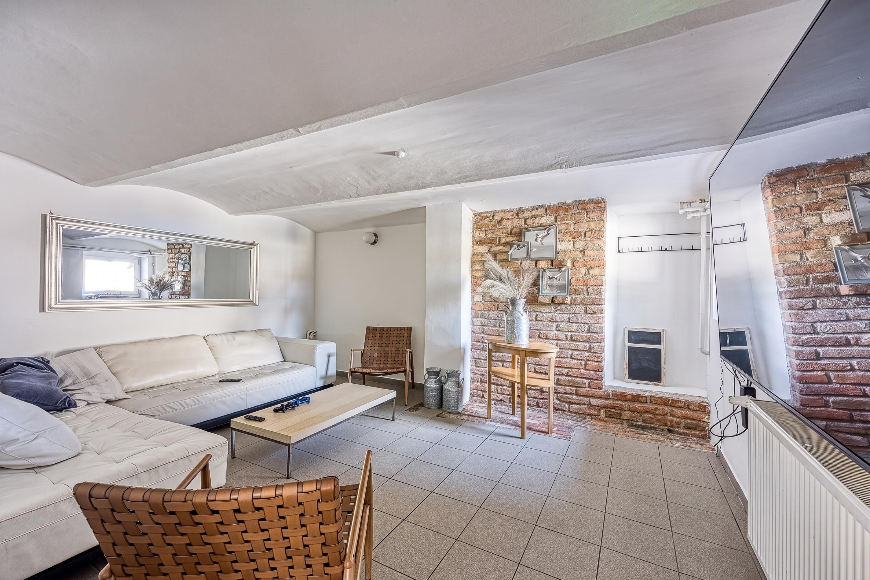 Ein Wohnzimmer mit einem weißen Sofa, einem Holztisch und einem Korbstuhl. Eine Ziegelwand mit einem Fenster und einem Spiegel ist zu sehen.