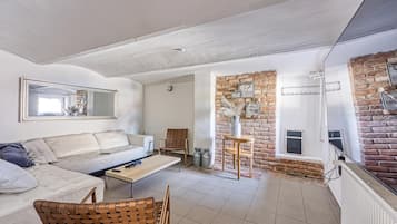 Ein Wohnzimmer mit einem weißen Sofa, einem Holztisch und einem Korbstuhl. Eine Ziegelwand mit einem Fenster und einem Spiegel ist zu sehen.
