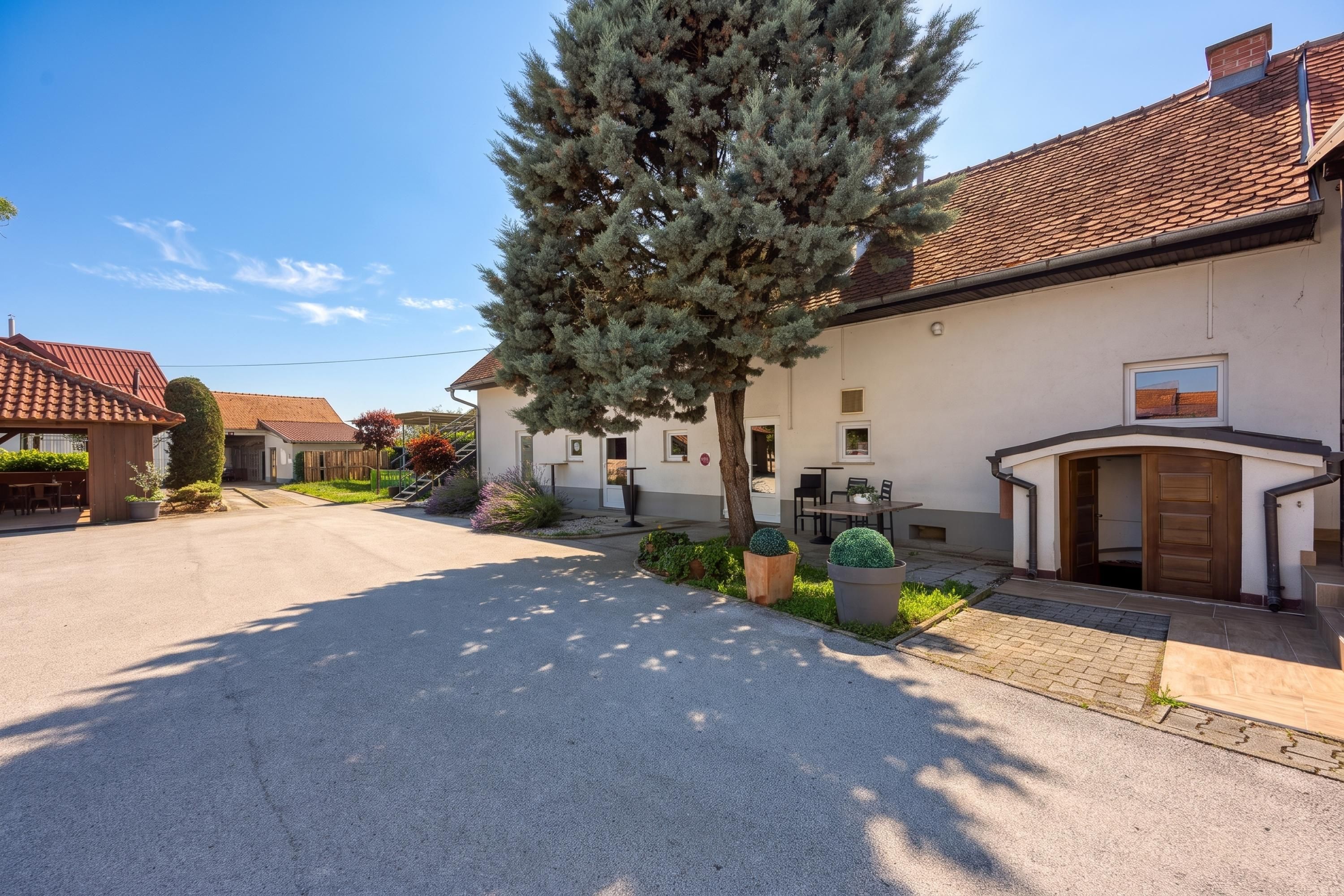 Ein Wohnhaus mit gepflasterter Auffahrt, einem großen immergrünen Baum und einem Gebäude mit einem Ziegeldach und einer kleinen Terrasse.