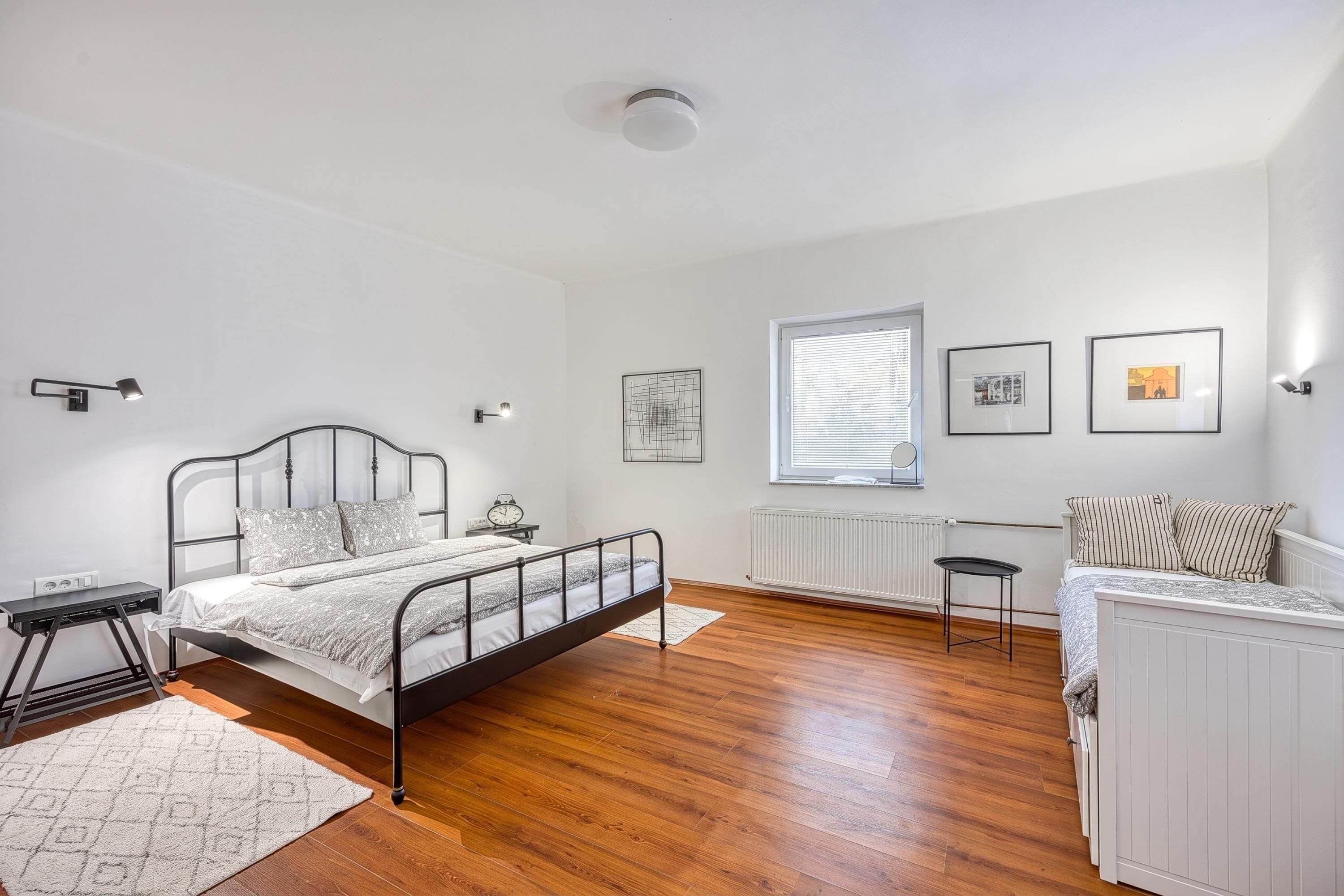 Ein Schlafzimmer mit Bett, Nachttischen, einem Heizkörper, einem Fenster mit Jalousien und Wandbildern.