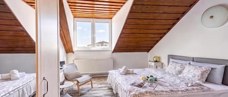 A cozy attic bedroom with a sloped ceiling, two beds, a chair, and a window with a view.