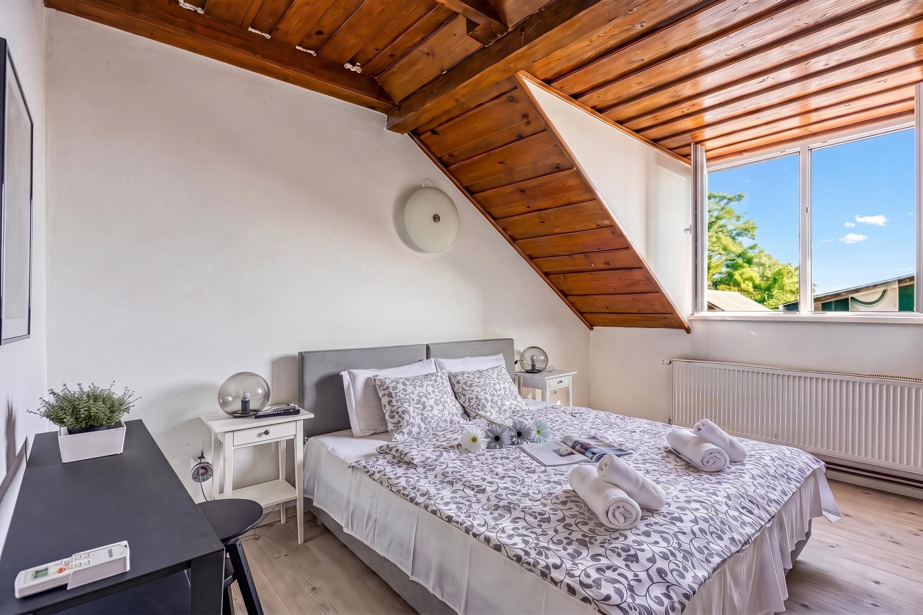 A bedroom with a sloped ceiling, a bed with patterned bedding, a bedside table with a lamp, and a window with a view of greenery.