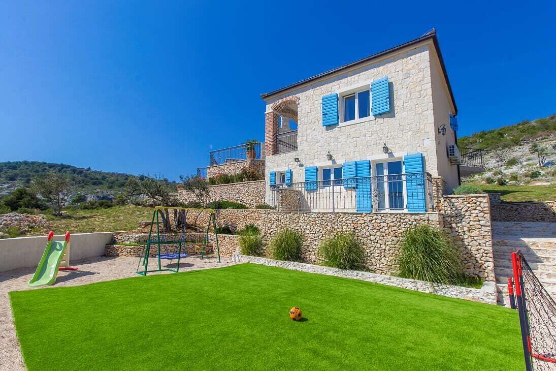 A two-story stone house with blue shutters, a small balcony, and a well-maintained lawn with a children's play area.