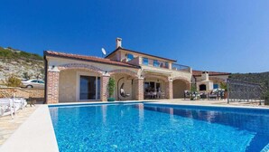 Una casa de dos pisos con piscina, un patio con mobiliario exterior y un paisaje montañoso de fondo.