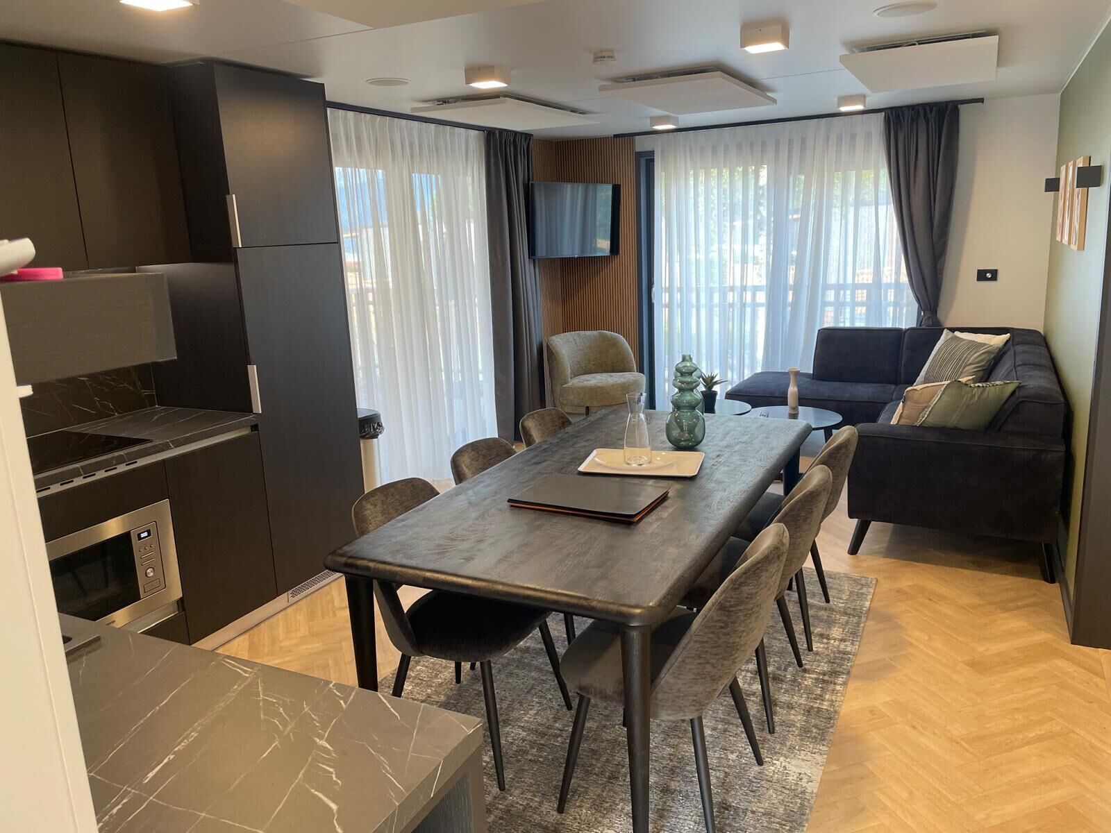 A modern kitchen with dark cabinetry, a built-in oven, and a marble countertop. There is a dining area with a long table and chairs, a sofa, and a TV mounted on the wall.