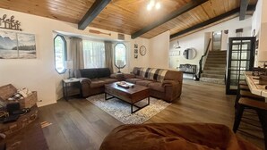 A cozy living room with a wooden ceiling, a large window with curtains, a sofa, a coffee table, and a staircase leading to another level. - 1 Mile to Summit or Village*Spa/Arcades/Pool Table (Big Bear Lake)