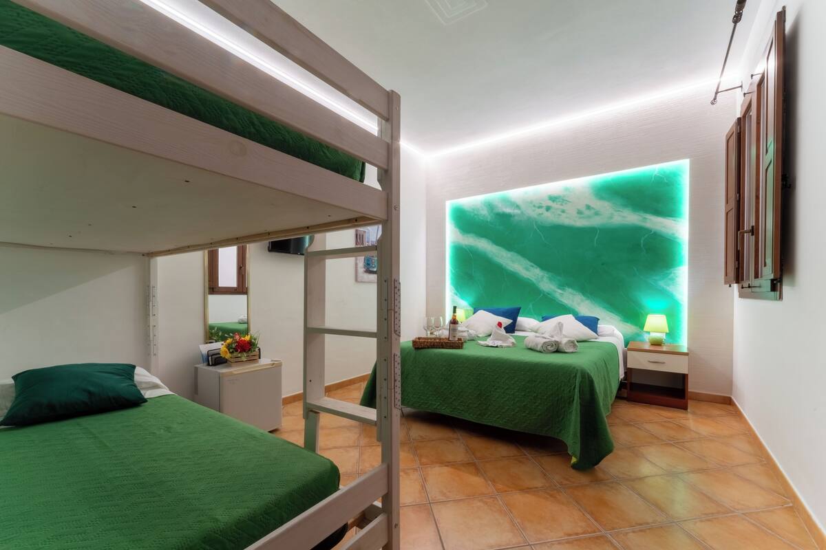 A room with bunk beds, a nightstand with a lamp, a wall-mounted mirror, and a green bedspread.