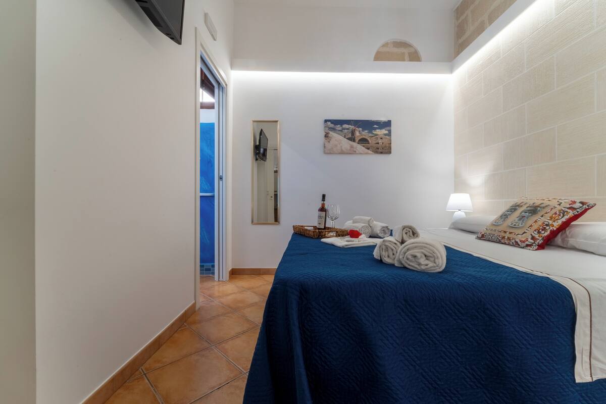 A hotel room with a bed, a nightstand, a lamp, a painting, and a door leading to another room.