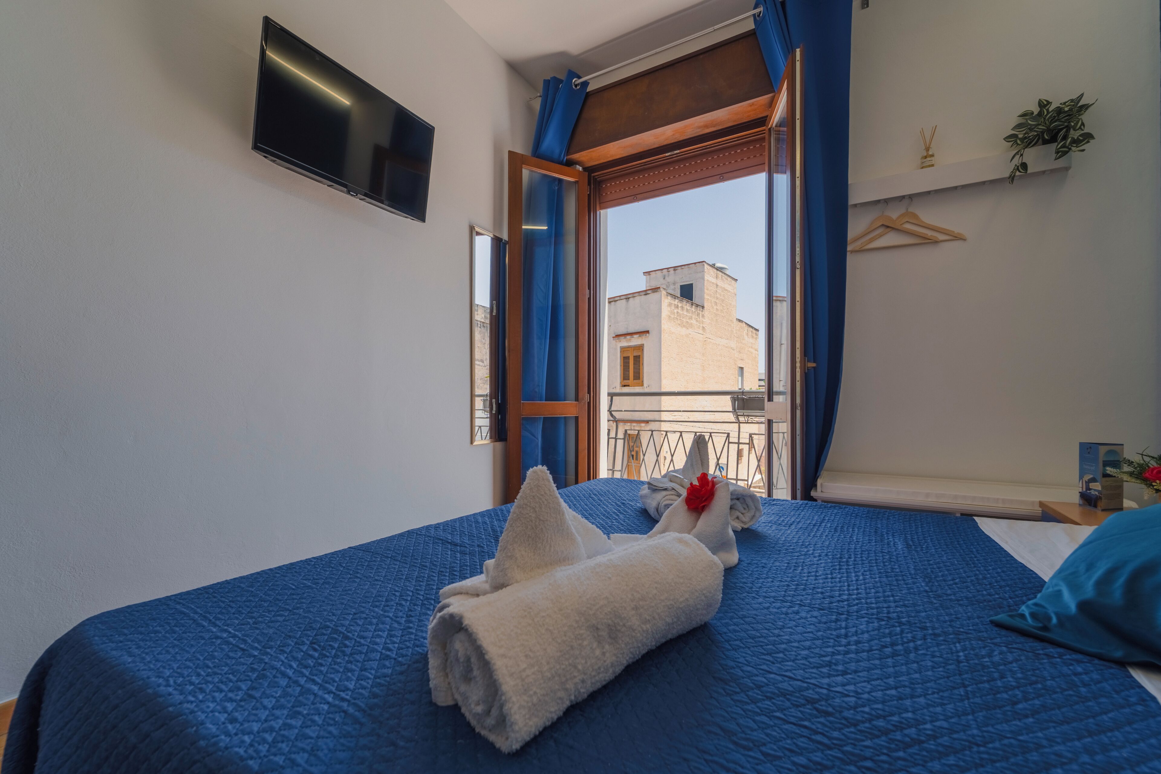 Ein Schlafzimmer mit einem Bett, einem an der Wand befestigten Fernseher, einem Fenster mit Vorhängen und Blick auf umliegende Gebäude.