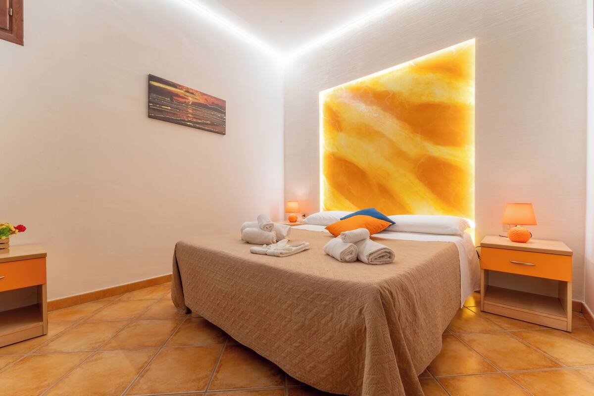 A hotel room with a bed, a nightstand, a lamp, a wall art piece, and a tiled floor.