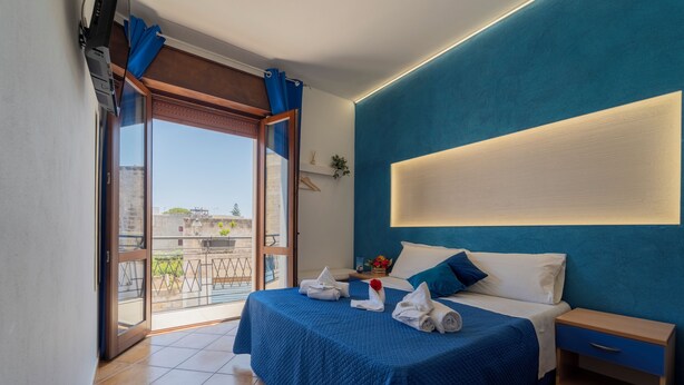 A bedroom with a bed, a nightstand, a window with curtains, and a view of buildings outside. - L'Oasi di Favignana Room and Charme (Favignana)
