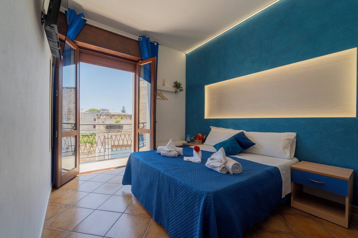 A bedroom with a bed, a nightstand, a window with curtains, and a view of buildings outside.