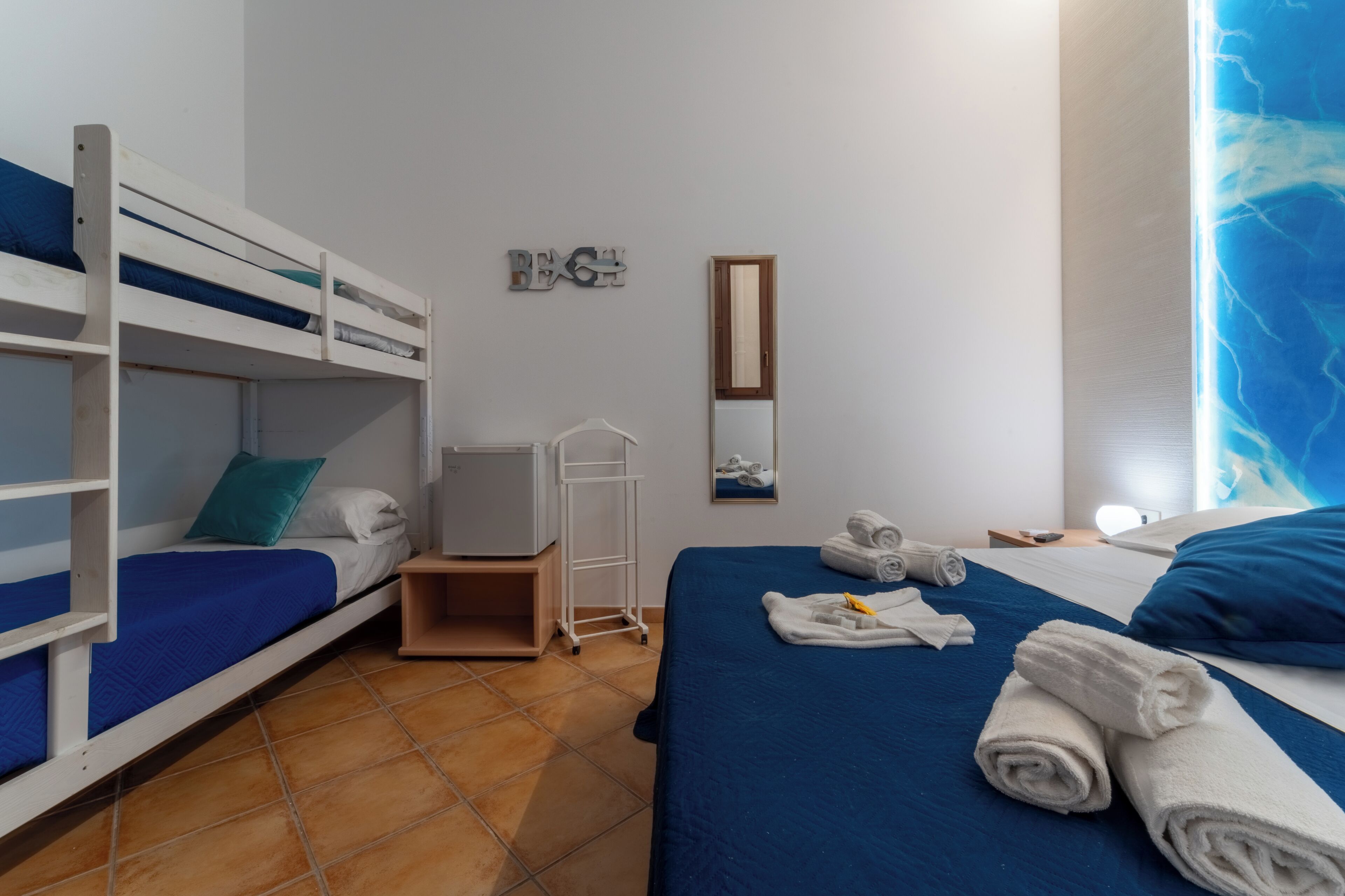 A room with bunk beds, a bed with rolled towels, a mirror, and a wall with a 'BEACH' sign.