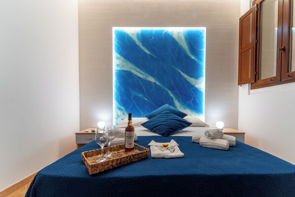 A neatly made bed with a blue bedspread, a bottle of wine, glasses, and towels on a bedside table.