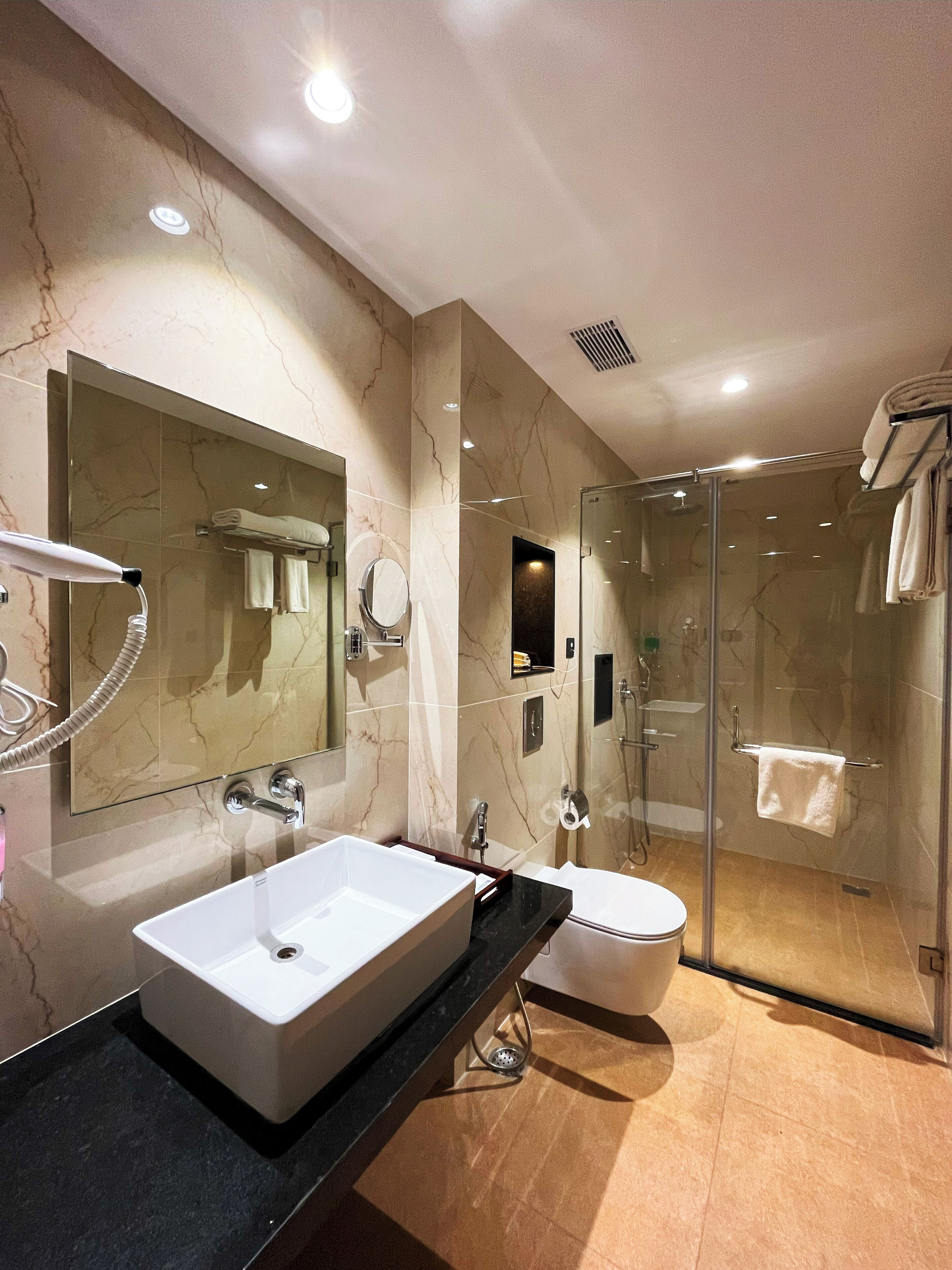A modern bathroom with a large mirror, two sinks, a glass shower enclosure, and marble walls.