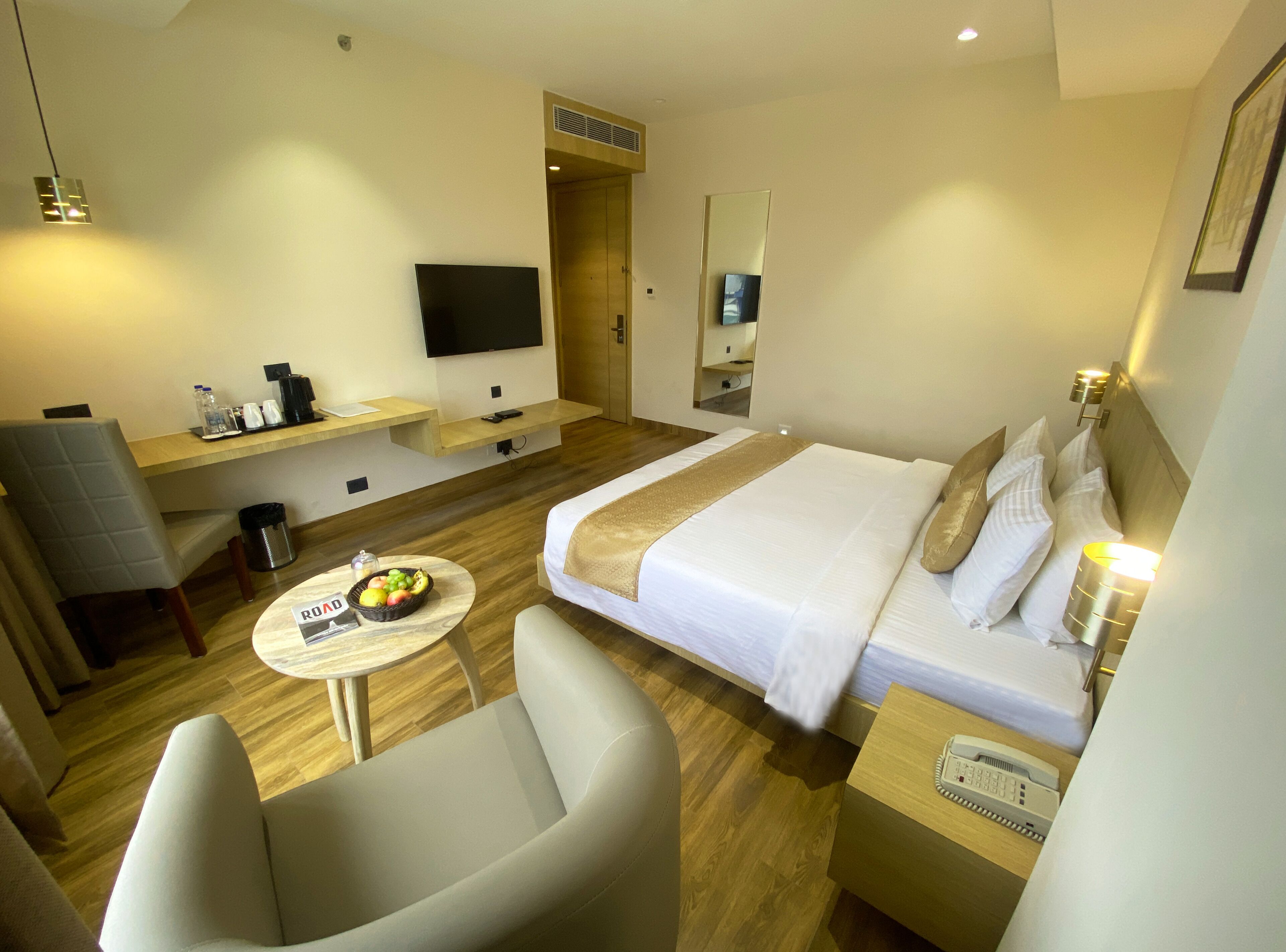 A hotel room with a bed, a desk, a chair, a small table with fruit, a trash bin, a phone, and a TV.