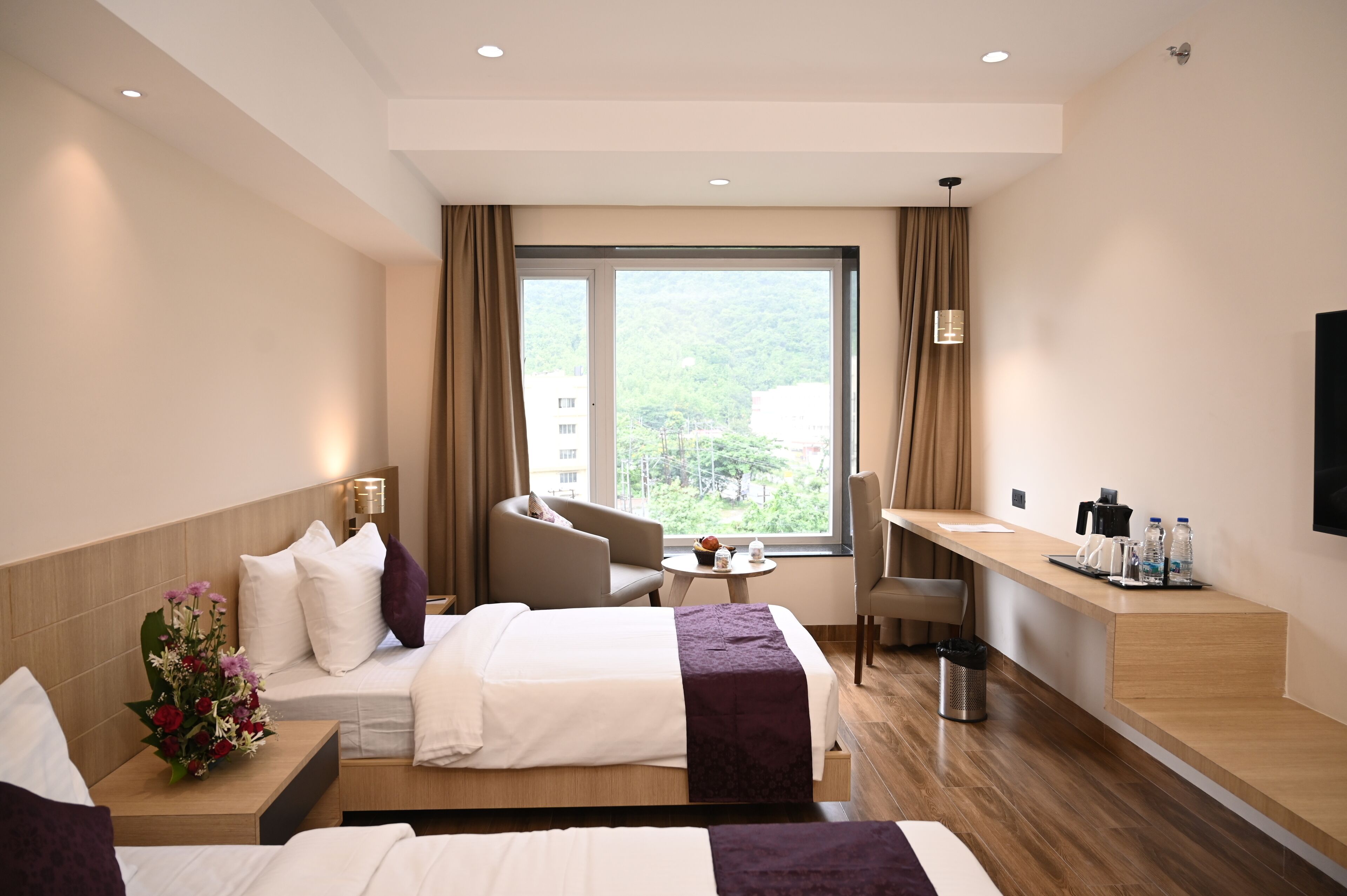 A hotel room with two beds, a desk, a chair, a small table, and a large window with a view of greenery.