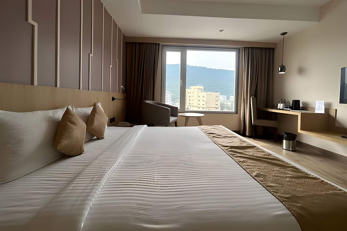 A hotel room with a large bed, a desk, a chair, and a view of the city and mountains.