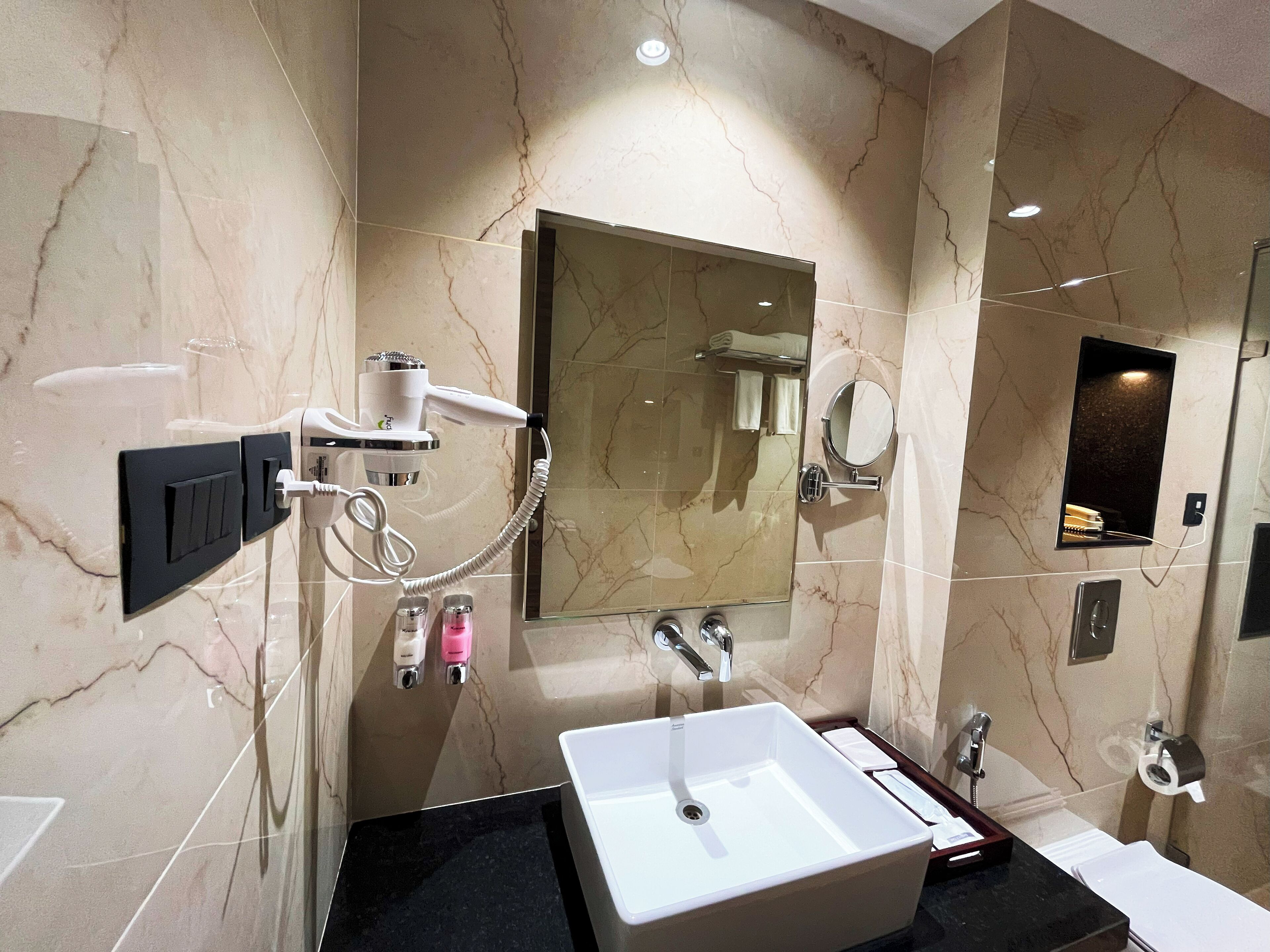Un baño moderno con un espejo grande, un lavabo blanco, un cabezal de ducha manual y un perchero para toallas.
