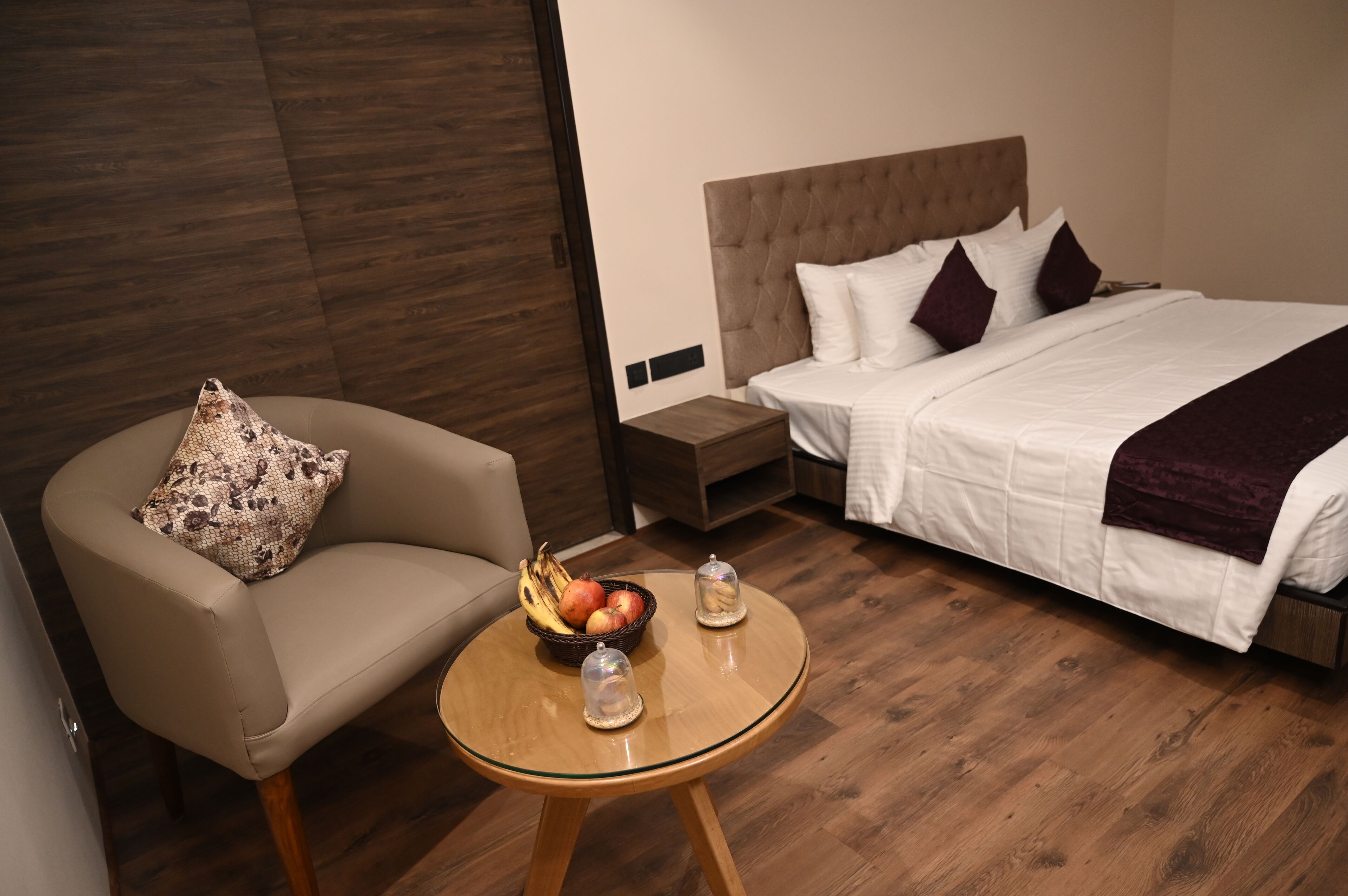 A hotel room with a bed, a chair, a small table with fruit, and a nightstand.