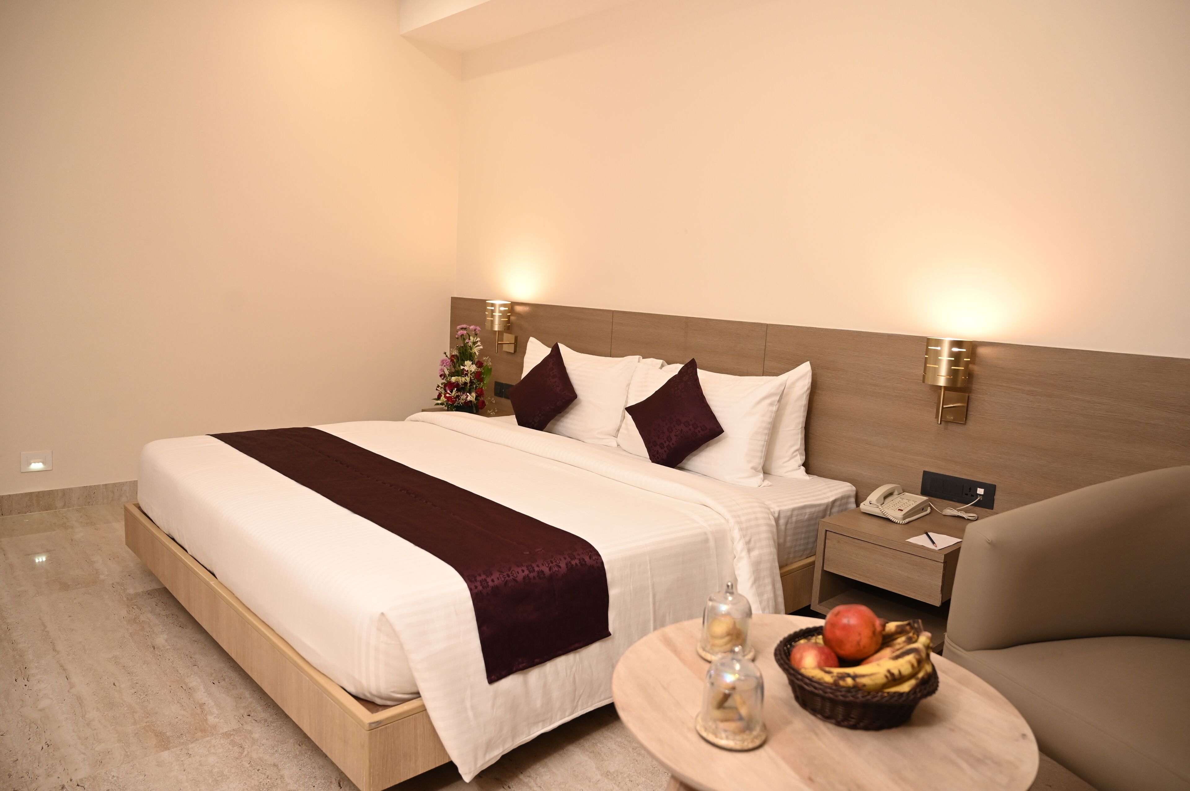 A hotel room with a large bed, bedside tables, a round table with fruits, and a wall-mounted phone.