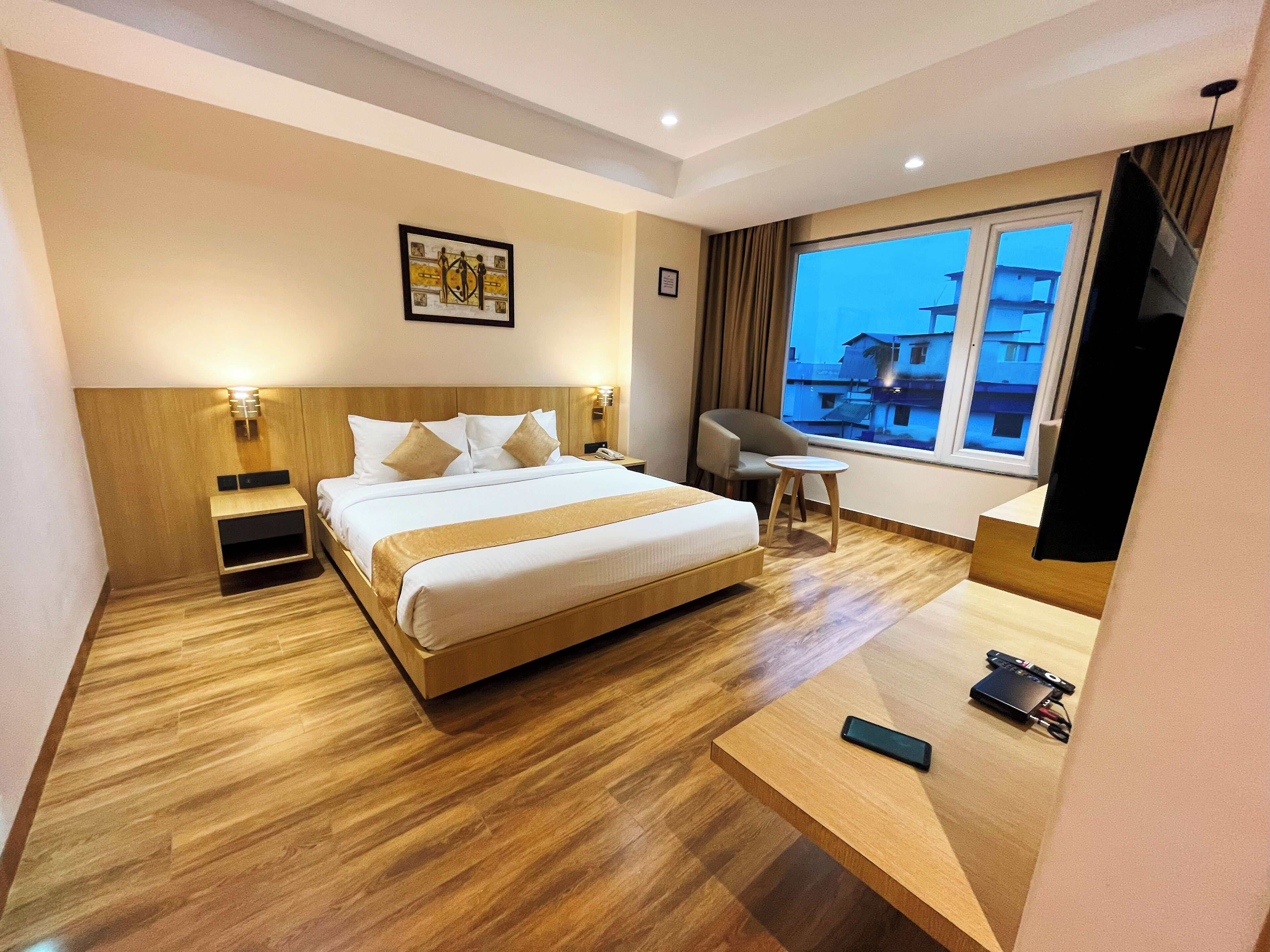 A hotel room with a large bed, bedside tables, a desk, a chair, a TV, and a window with a view of buildings.