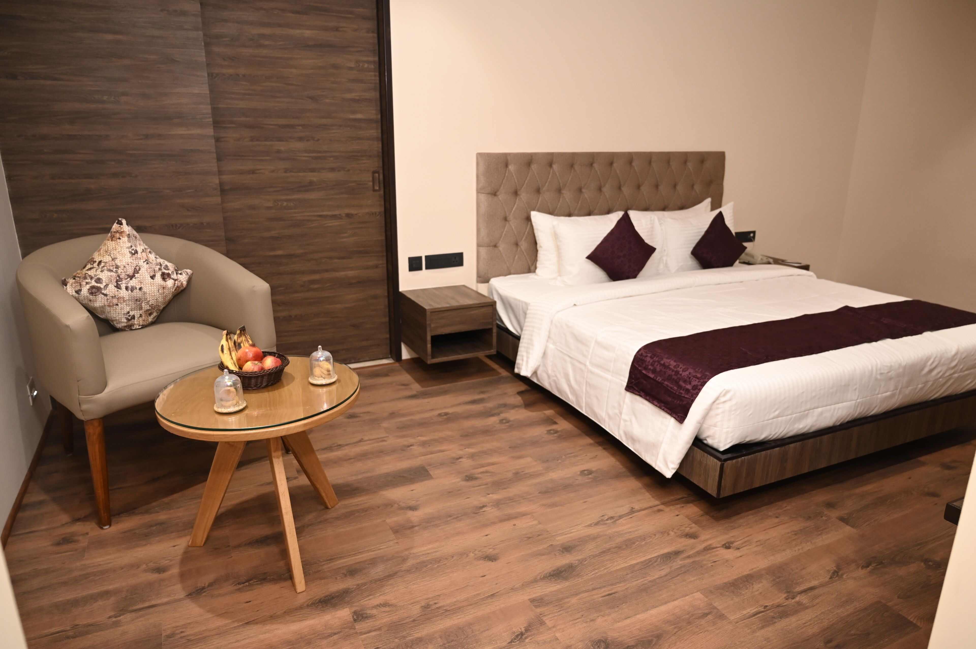 A hotel room with a bed, a chair, a small table with fruit, and a wooden floor.