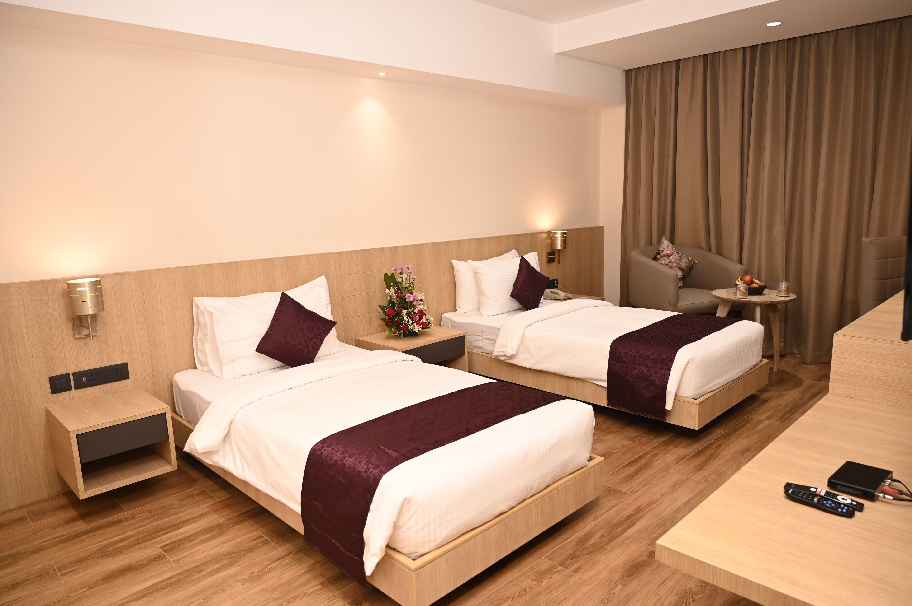 A hotel room with two beds, a desk, a chair, a nightstand, and a wall-mounted lamp.