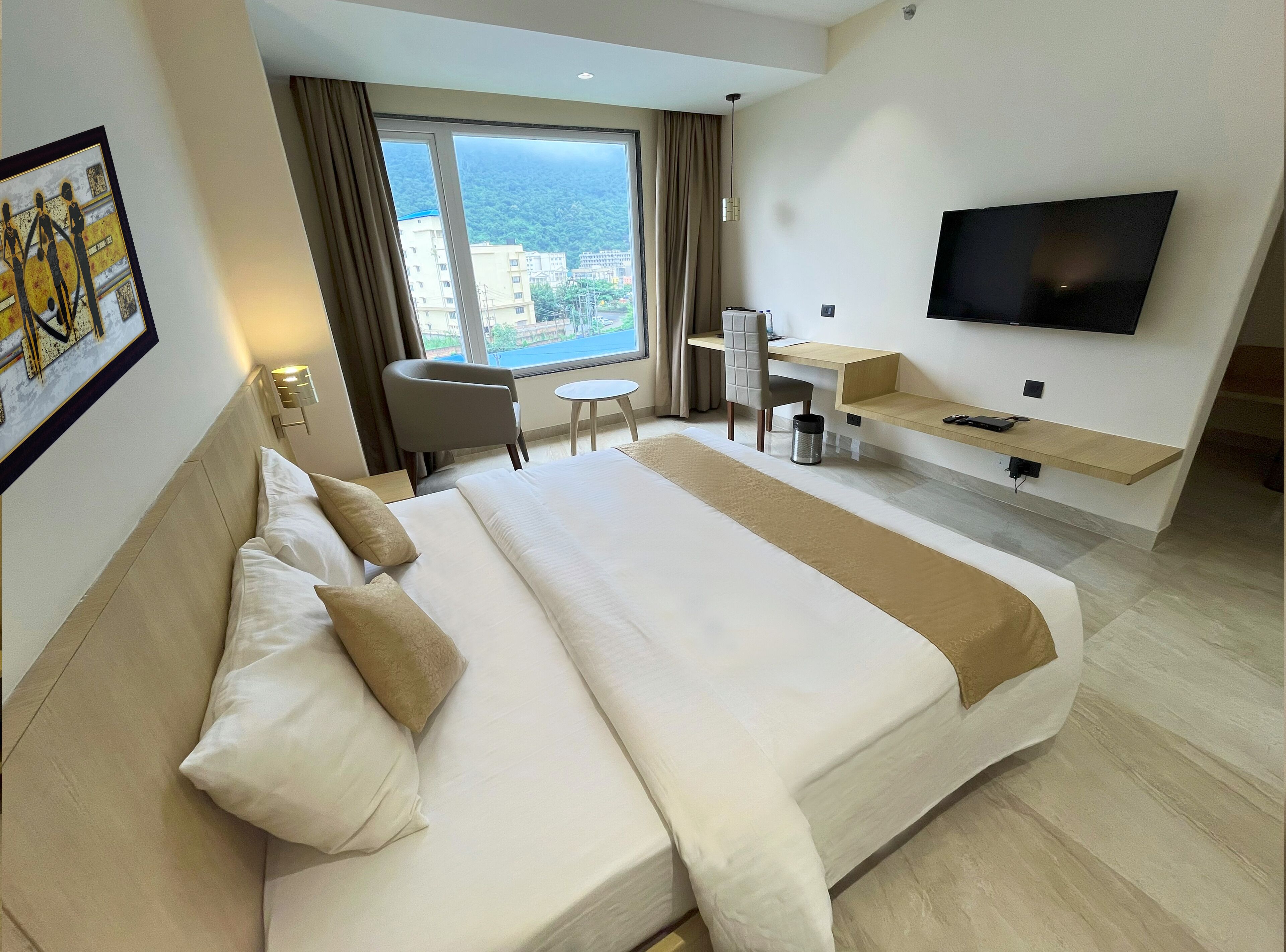 A hotel room with a large bed, a desk, a chair, a TV, and a view of the city.
